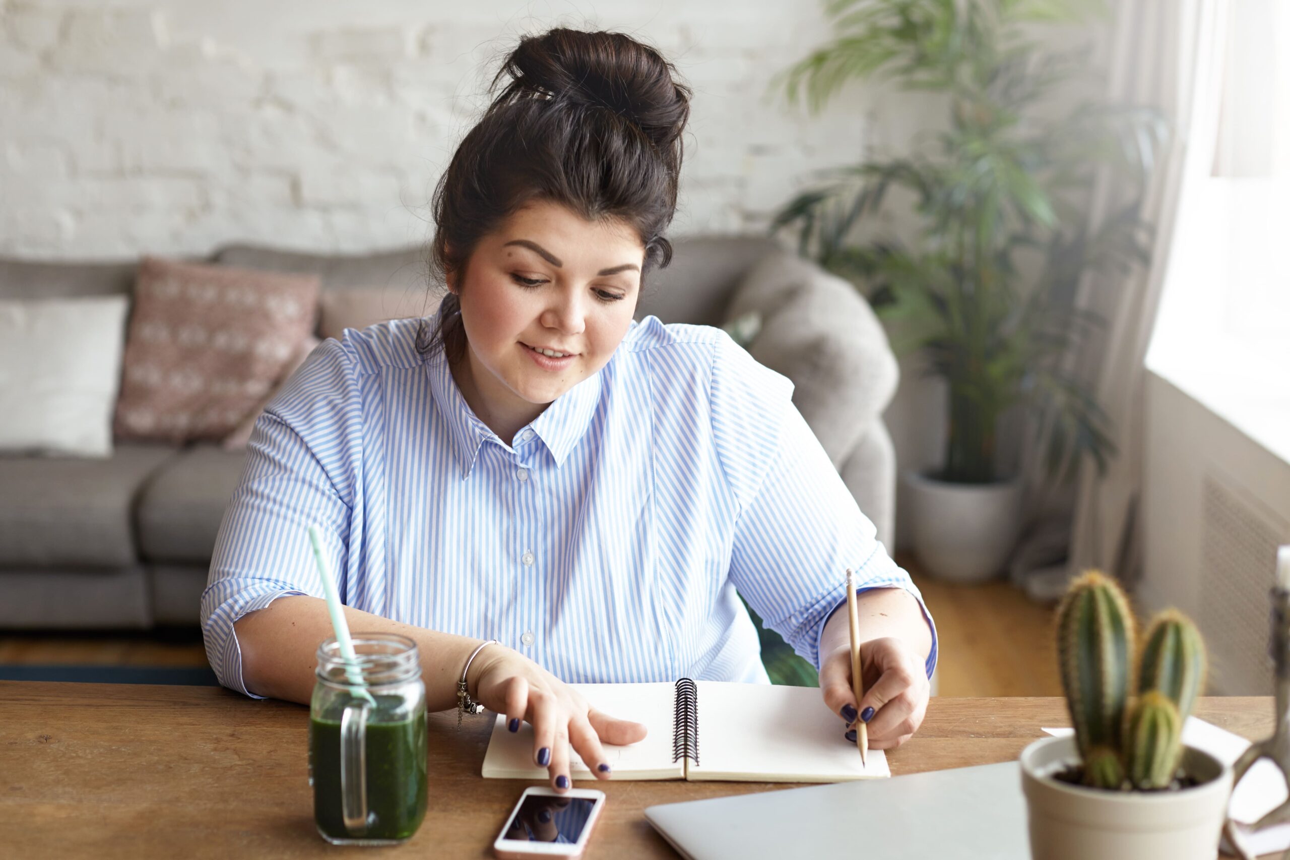 Smiling woman sitting at a bright wooden desk, writing in a notebook while looking at her phone, representing planning for virtual sober coach services. A green smoothie, laptop, and houseplant are also visible.