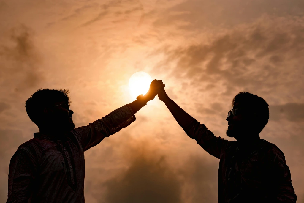 A silhouette of two people facing each other against a bright, hazy sunset, with their raised hands touching in the center to frame the sun, symbolizing connection and alcohol recovery.