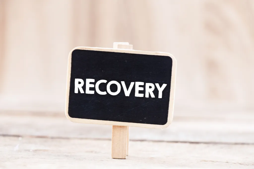A small black chalkboard sign with the word RECOVERY in bold white letters, held by a wooden clip against a soft, blurred background. An encouraging image representing the journey of addiction recovery.